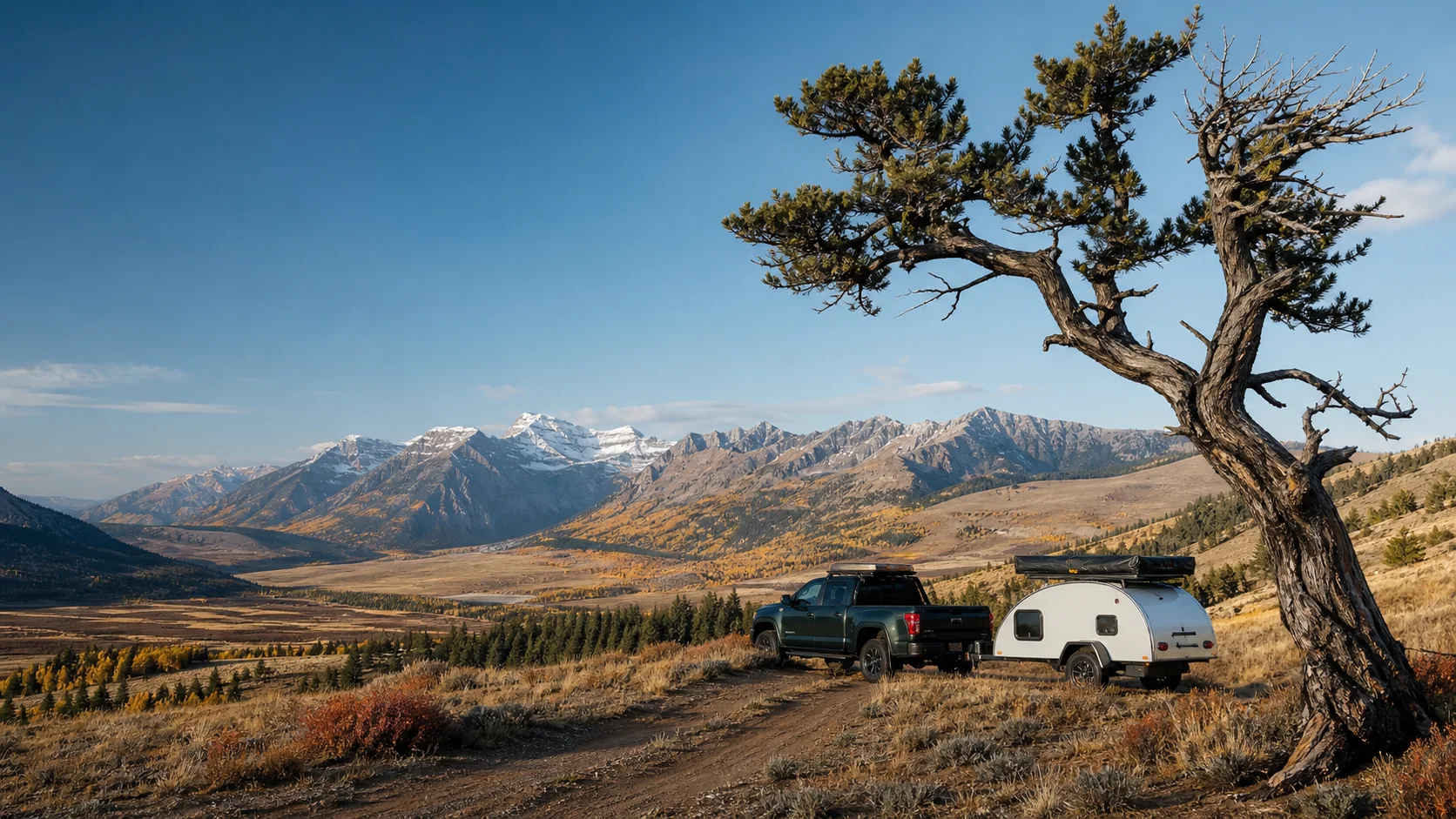 A pickup and teardrop trailer beneath a crooked tree in a wide autumn mountain valley